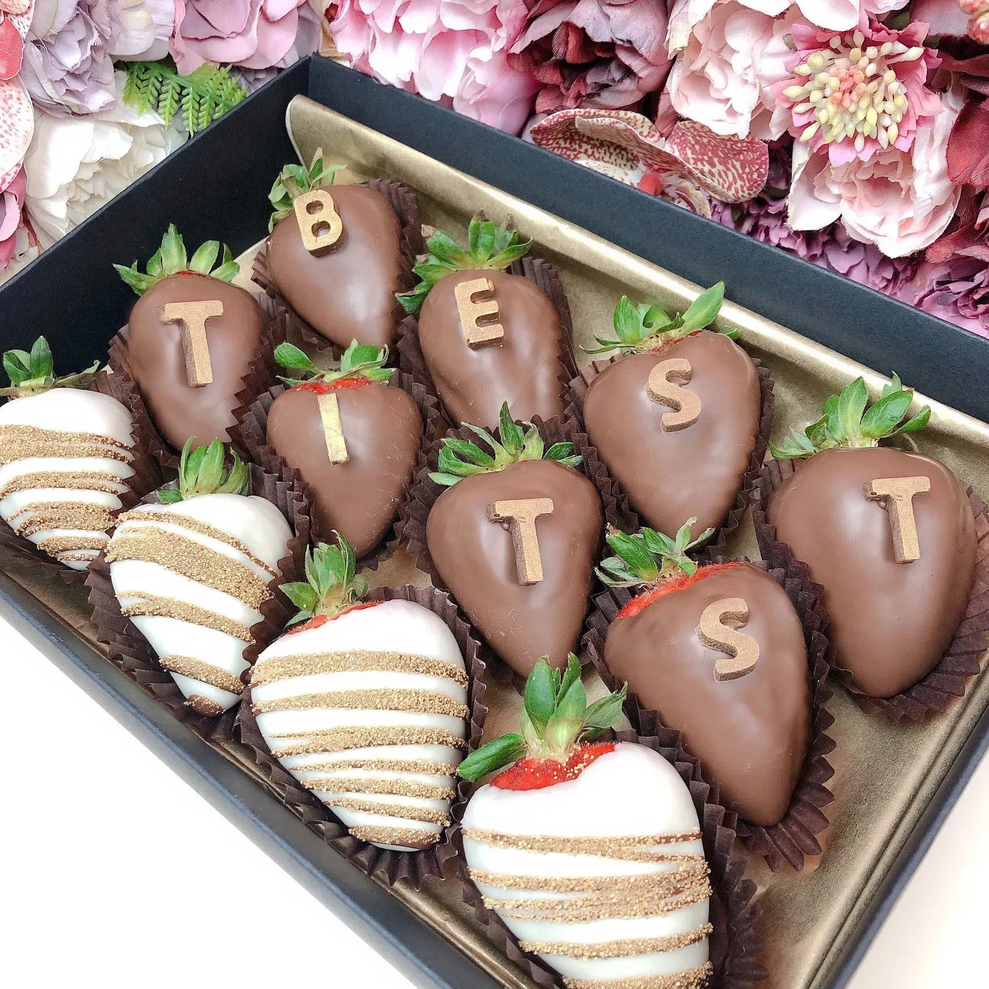 A box of large strawberries covered in chocolate. Some are coated in milk chocolate with chocolate letters on top, while others are dipped in white chocolate with gold stripes. The box is perfect as a girlfriend naughty gift