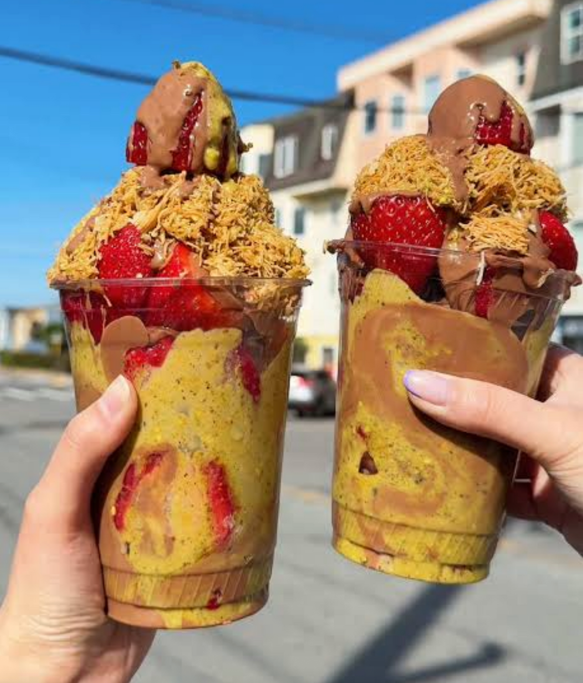 Close-up of an individual catering cup filled with fresh sliced strawberries, thick premium milk chocolate glaze, and toasted golden kataifi pastry mixed with green pistachio paste.